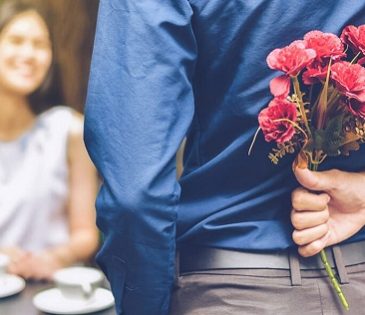 man holds flowers behind his back while approaching a woman