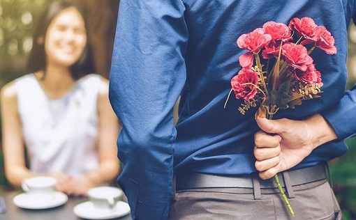 man holds flowers behind his back while approaching a woman