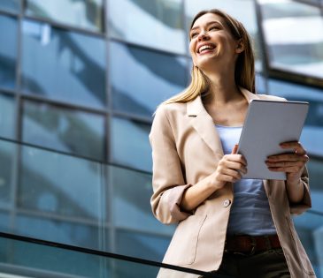 smiling blonde woman in a taupe colored blazer holding an ipad